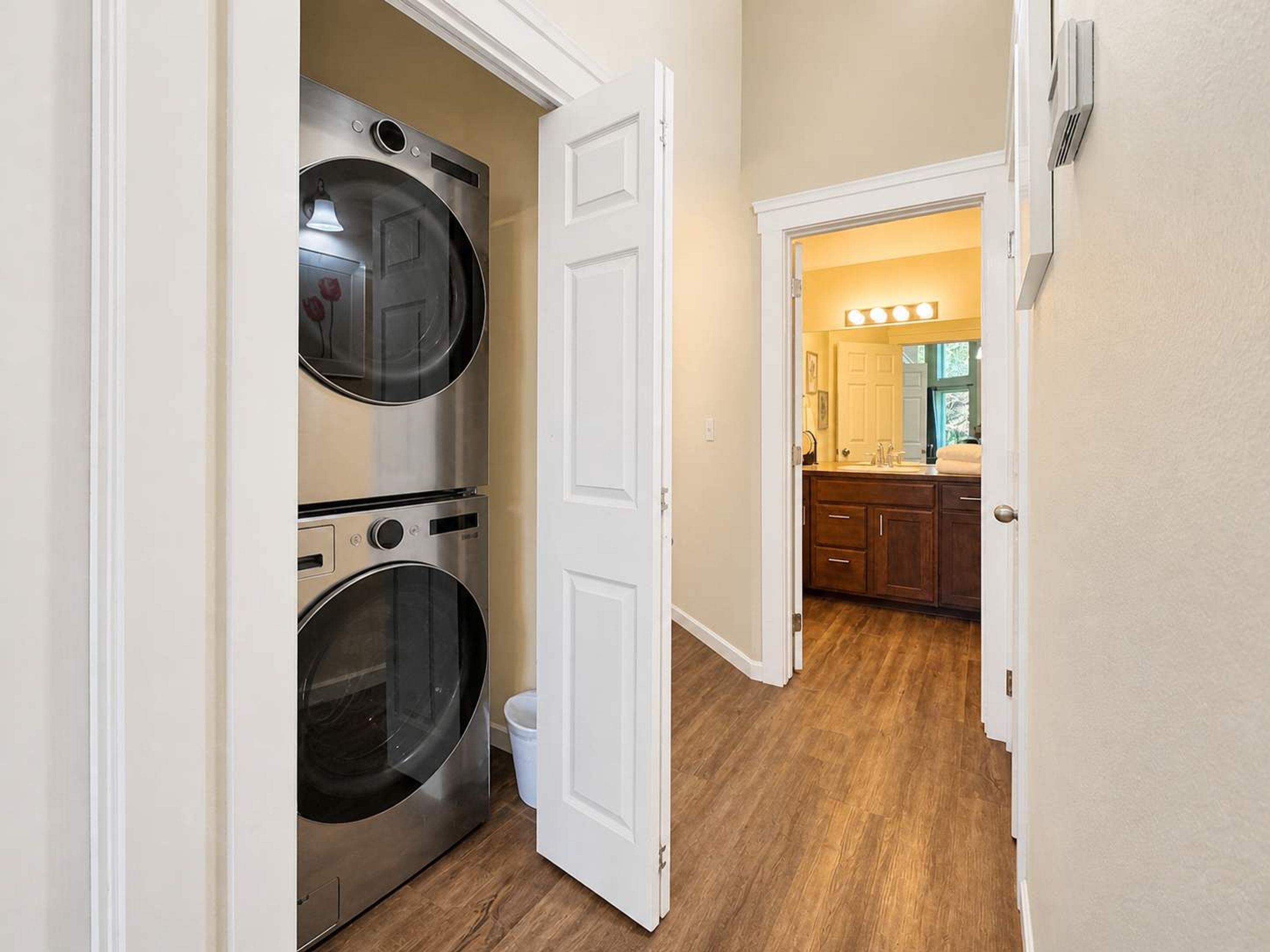 Un cuarto de lavado con lavadoras y secadoras apiladas, una puerta blanca que da a una cocina con armarios de madera y un lavamanos.