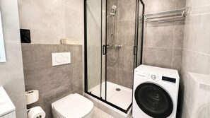 A modern bathroom with a glass-enclosed shower, a white toilet, and a front-loading washing machine. - 1 room Apartment with AC, Vienna Gate 3 (Bratislava)