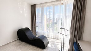Una sala de estar moderna con un sillón negro, un ventanal con cortinas translúcidas y vistas a edificios.