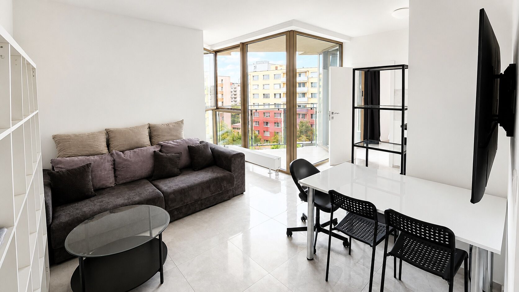 A modern living space with a sofa, a dining table, and chairs, a TV mounted on the wall, and a large window offering a view of buildings.