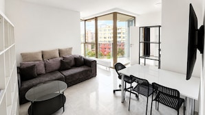 En moderne stue med sofa, spisebord og stole, et fjernsyn ophængt på væggen og et stort vindue med udsigt til bygninger.