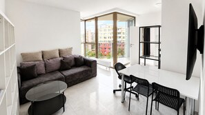 Un espace de vie moderne comprenant un canapé, une table à manger et des chaises, un téléviseur fixé au mur et une grande fenêtre donnant sur des immeubles.