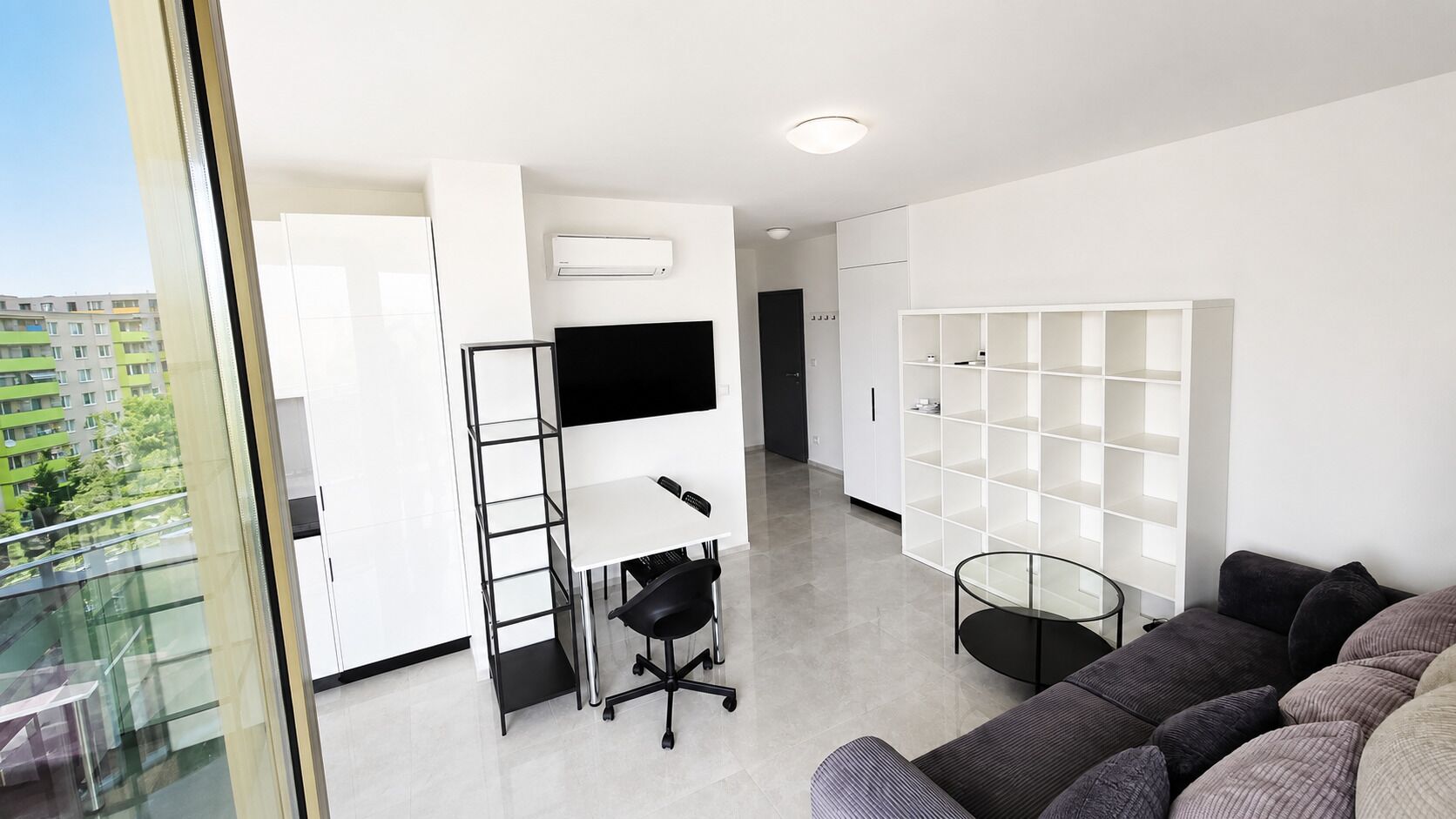 Una sala de estar moderna con un sofá, una televisión, una estantería blanca y un ventanal que da a un balcón.