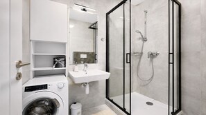 A modern bathroom with a glass-enclosed shower, a built-in washing machine, and a white sink. - 2 room Apartment with AC, Vienna Gate 4 (Bratislava)