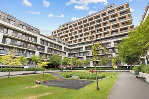 A modern apartment complex with multiple balconies, a central courtyard with grass and trees, and a pedestrian pathway.