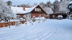 A snow-covered residential area with a wooden house, a garage, and a vehicle parked outside. - Apartment 'Jagdhaus An Der Nemitzer Heide' with Wi-Fi (Prezelle)