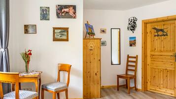 Une pièce avec un plancher en bois, une porte en bois et un meuble en bois orné d’un motif représentant une empreinte de patte de chien. On y trouve deux chaises en bois, dont une avec un coussin et l’autre sans. Une petite table avec un vase de fleurs est visible.