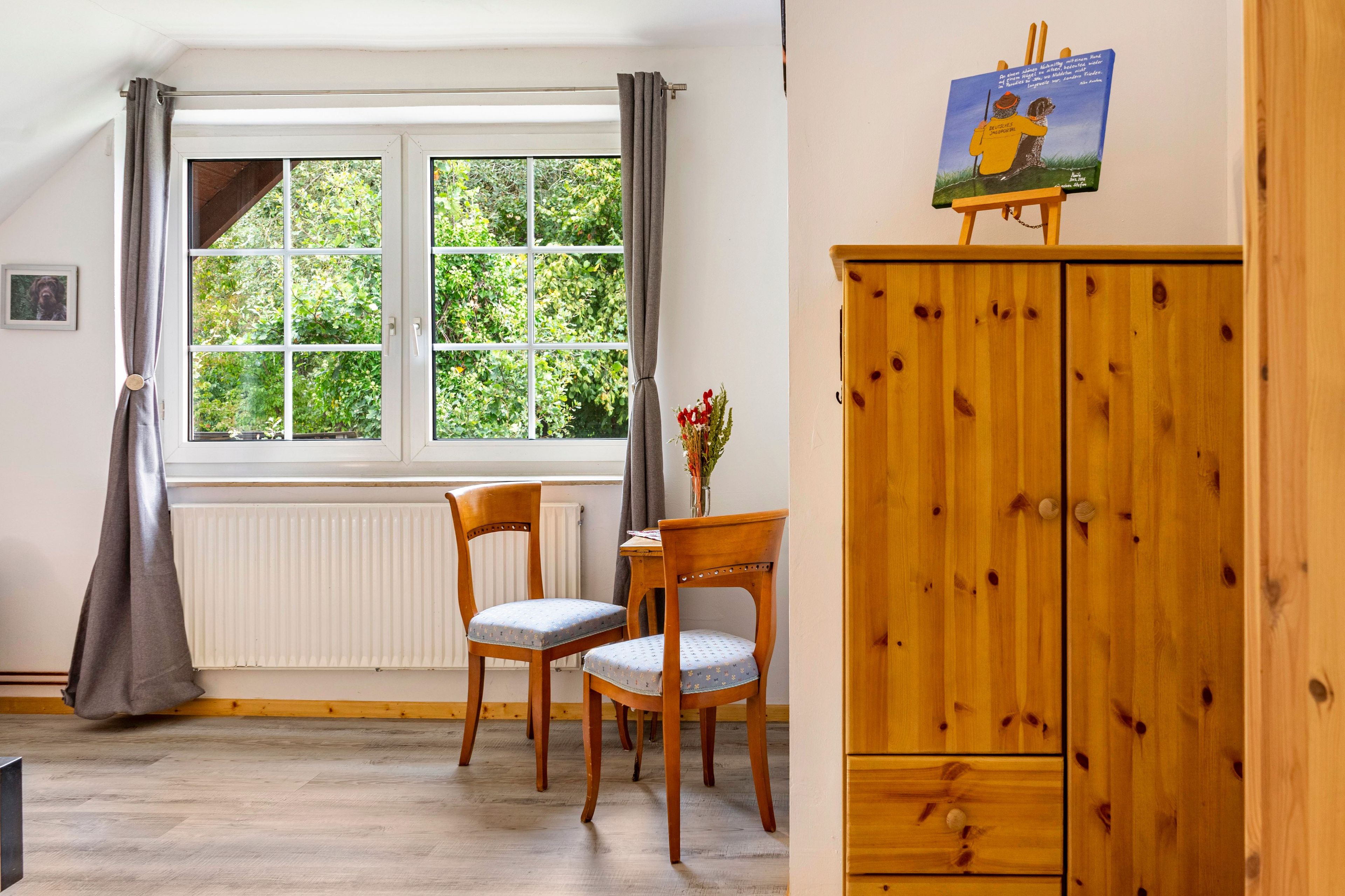 Een kamer met een raam, twee stoelen, een houten kast en een schilderij aan de muur.