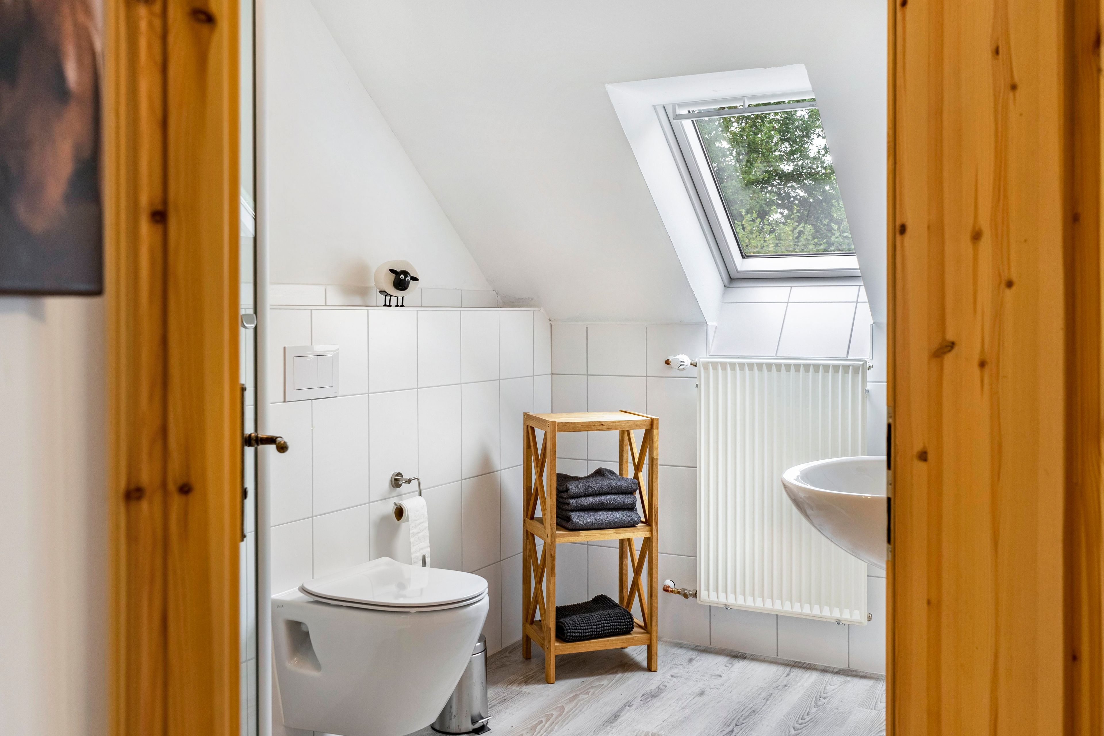 A modern bathroom with a skylight, white tiles, a toilet, a wooden shelf with towels, and a radiator.