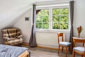 A room with a large window, a fur chair, a wooden chair, a small table with a vase of flowers, and a radiator.