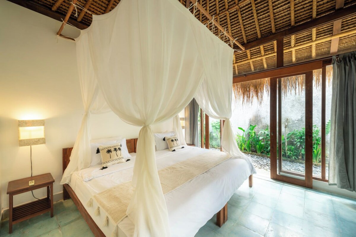 A bed with a canopy, a bedside table, and a view of greenery outside.