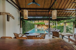 An outdoor pool area with a thatched roof, wooden furniture, and a view of a lush garden.
