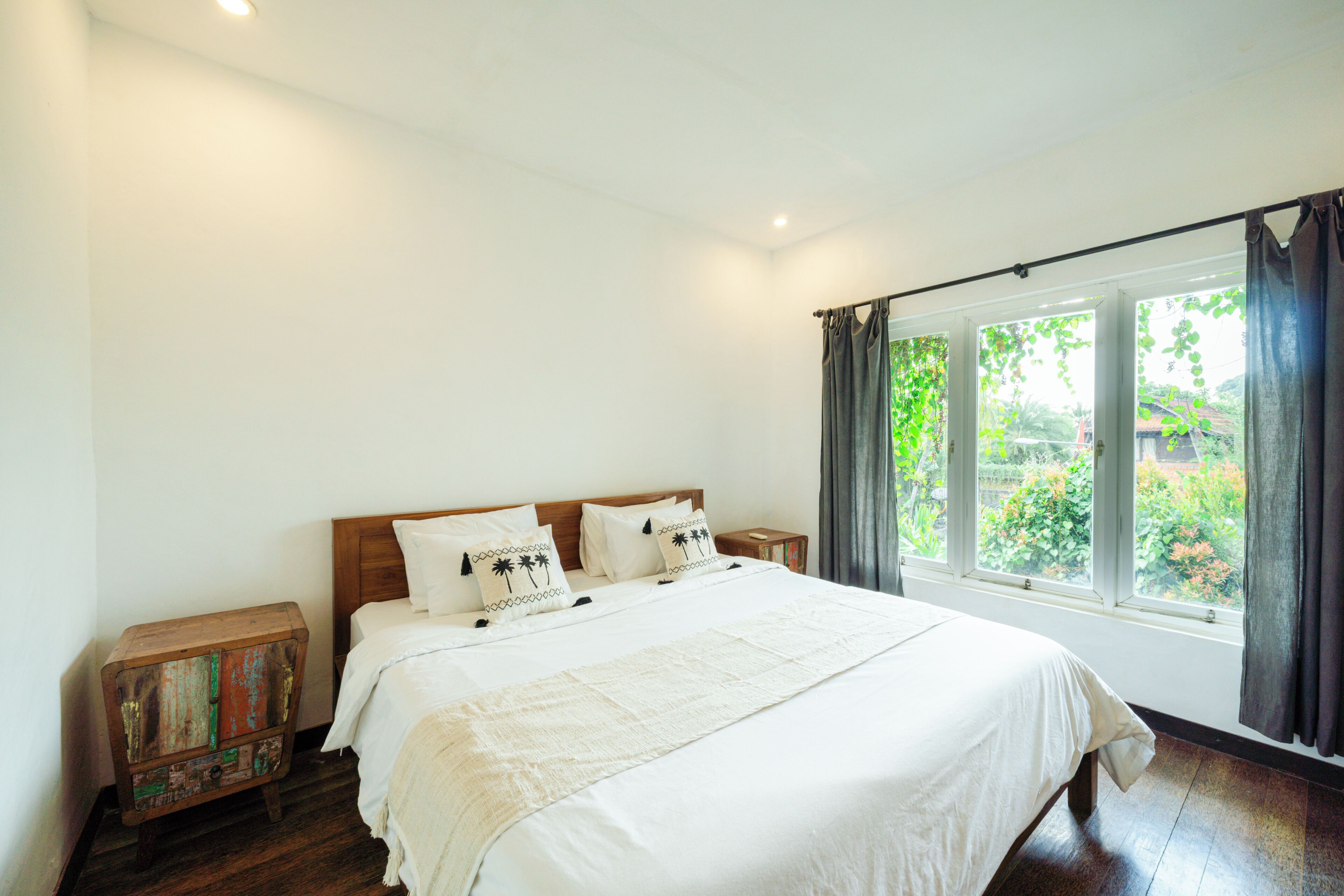 A bedroom with a wooden bed, white bedding, a small wooden nightstand, and a window with curtains.