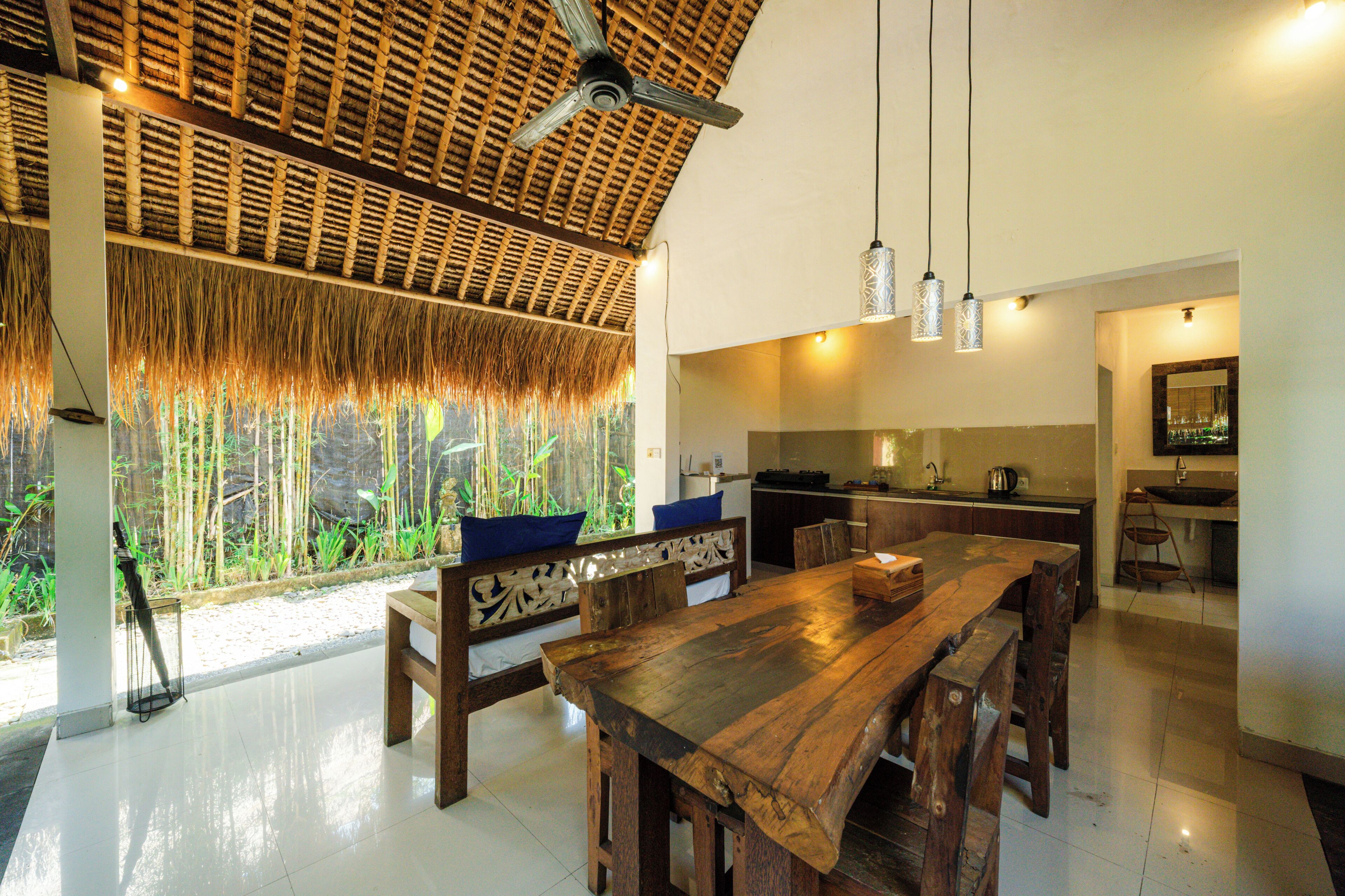 A spacious dining area with a large wooden table and benches, a kitchen with modern appliances, and a view of bamboo plants through the window.