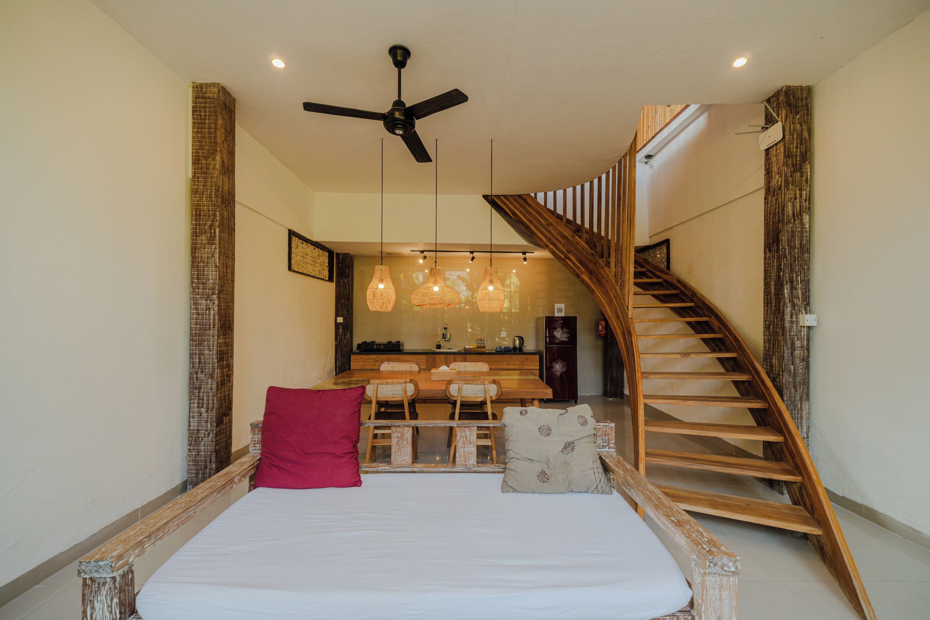 Ein Schlafzimmer mit einem Bett, einem roten Kissen, einem hölzernen Kopfende, einem Deckenventilator und einer Treppe zu einem Küchenbereich.