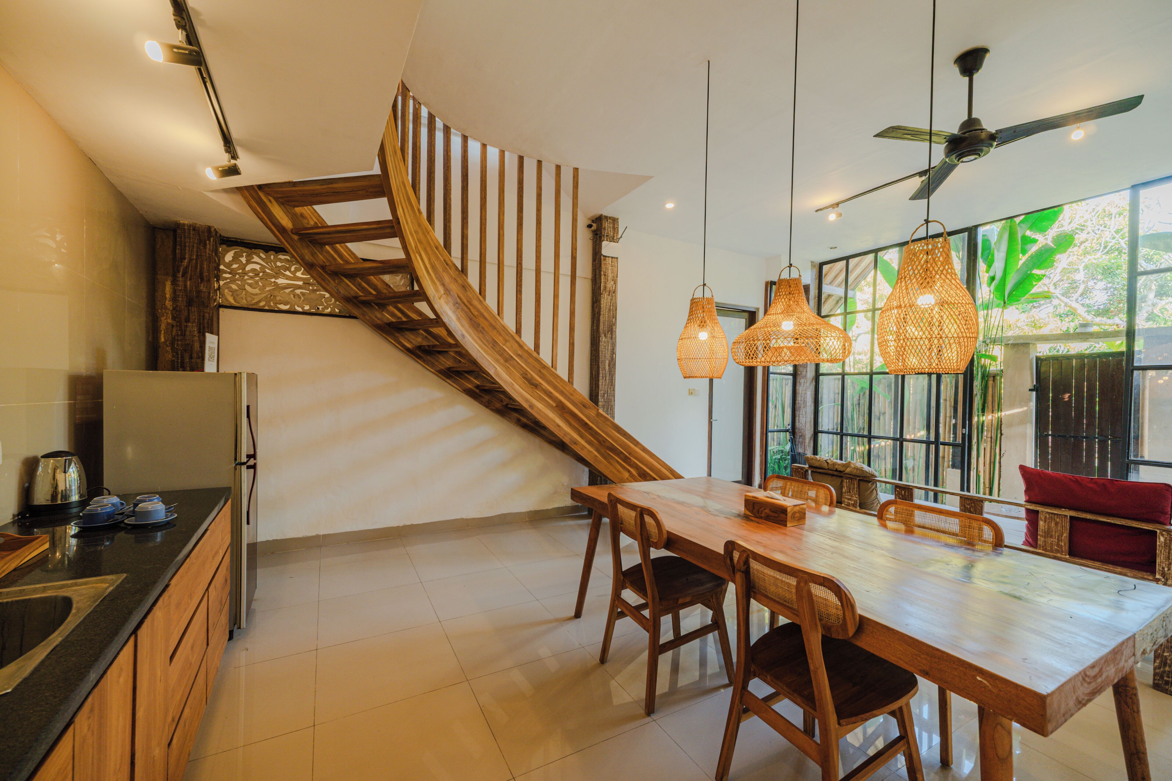 A modern kitchen with a wooden staircase, a dining area with a large table and chairs, and a ceiling fan.