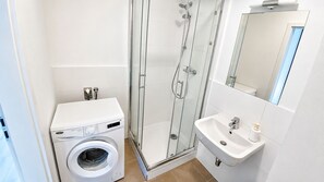 A compact bathroom with a glass-enclosed shower, a washing machine, and a sink. - 1 room apartment, balcony, Zuzany Chalupovej 18 (Bratislava)
