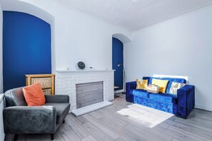 Uma sala de estar com lareira, sofá, poltrona e uma pequena mesa.