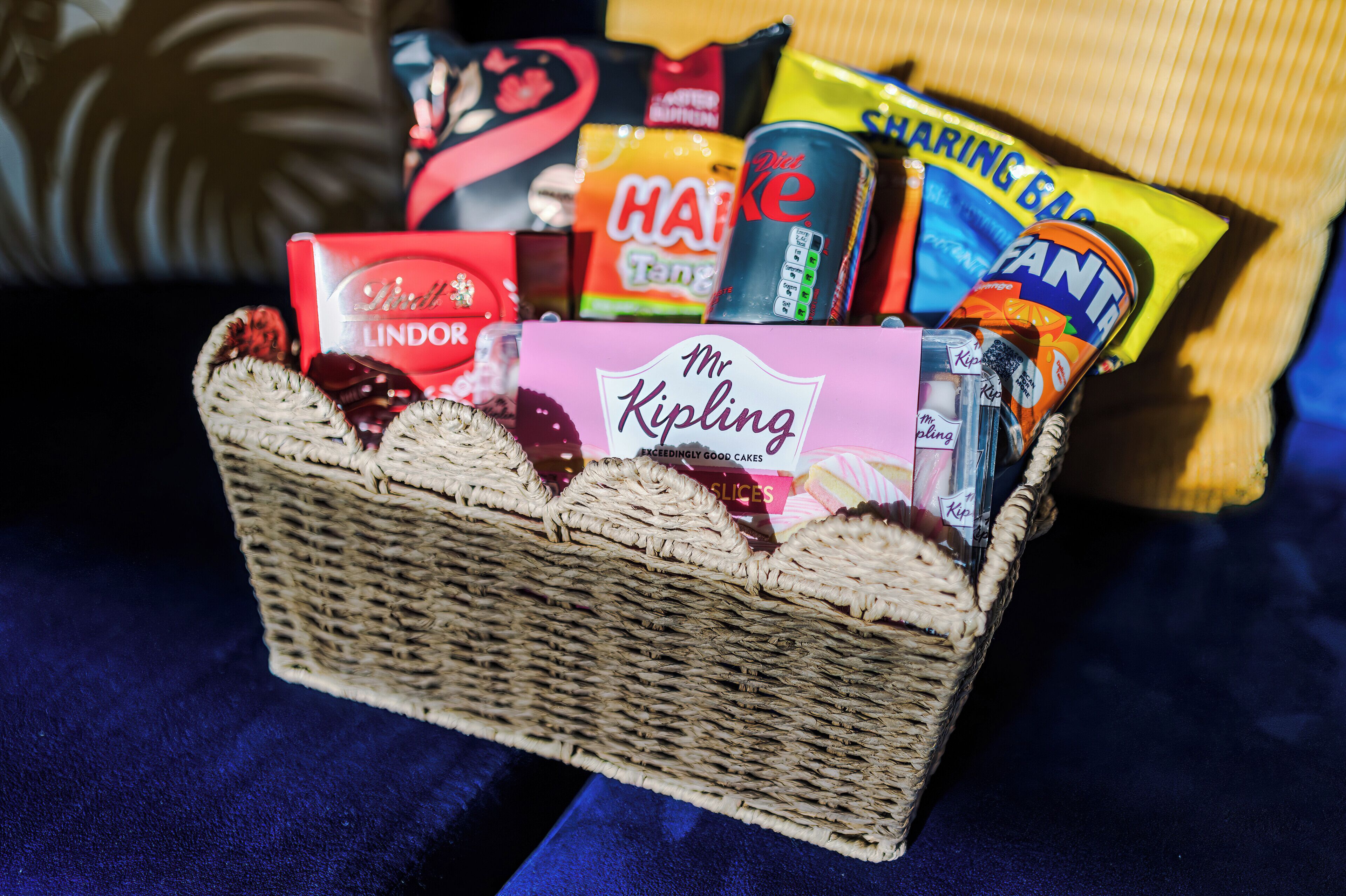 A wicker basket filled with various snack items including a can of diet coke, a pack of Haribo gummies, and a box of Lindor chocolates.