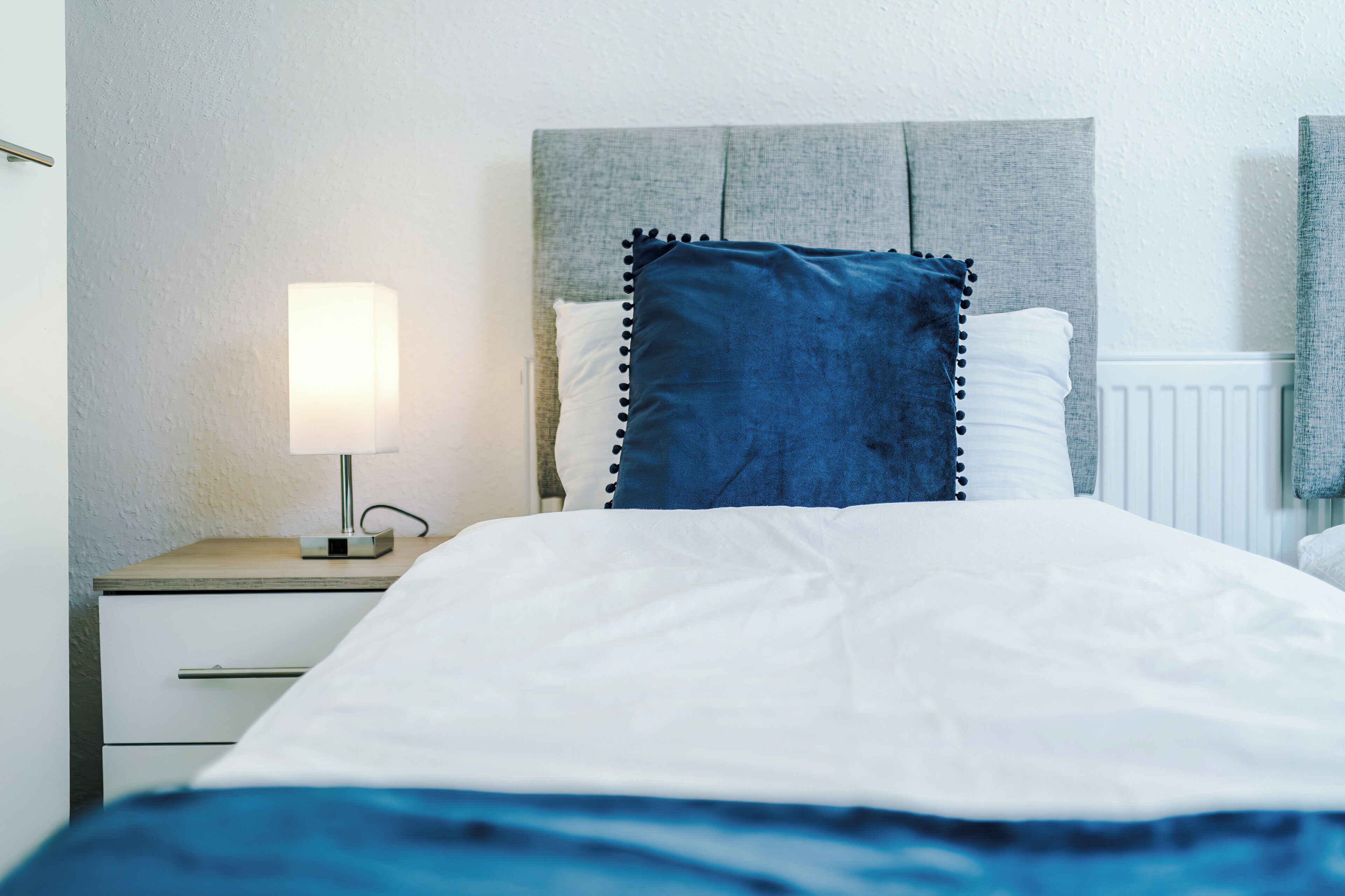 A neatly made bed with a white duvet and a blue accent pillow, a bedside lamp, and a wooden nightstand.