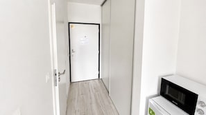 A narrow hallway with a white door at the end, a microwave on the right, and a wooden floor. - 1 room apartment, balcony, Zuzany Chalupovej 18B (Bratislava)
