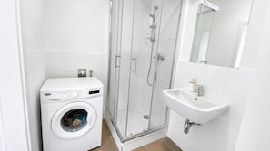 A compact bathroom with a washing machine, a glass-enclosed shower, and a sink. - 1 room apartment, balcony, Zuzany Chalupovej 18B (Bratislava)