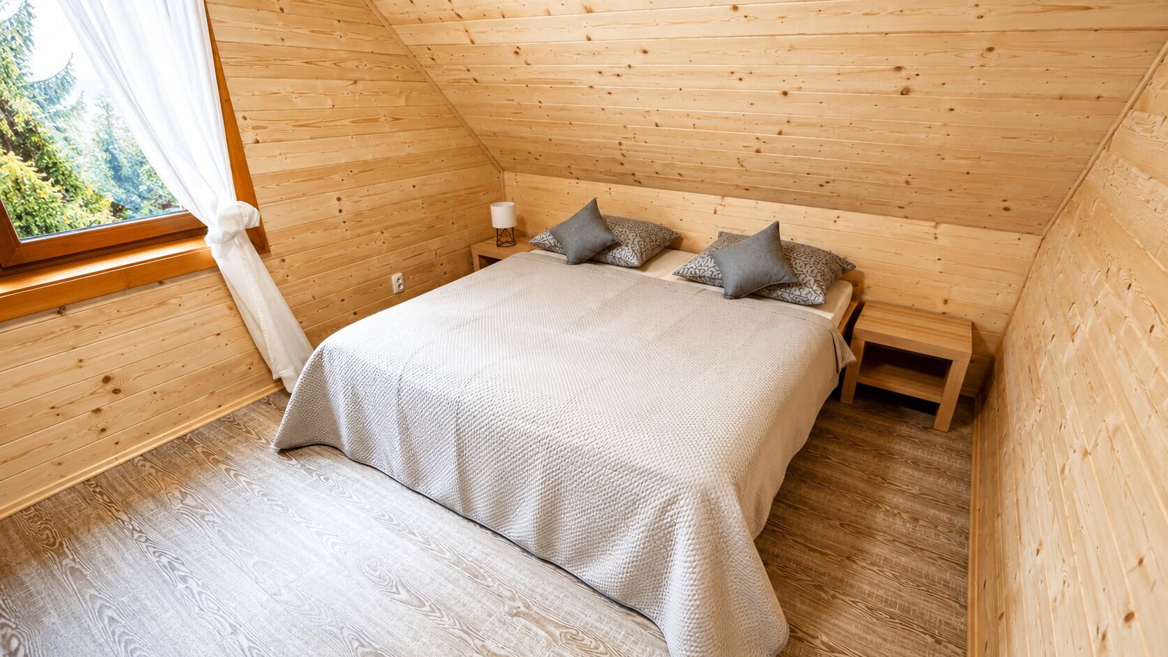 Une chambre dans une cabane en bois, avec un lit, des oreillers, une table de chevet et une fenêtre avec des rideaux.
