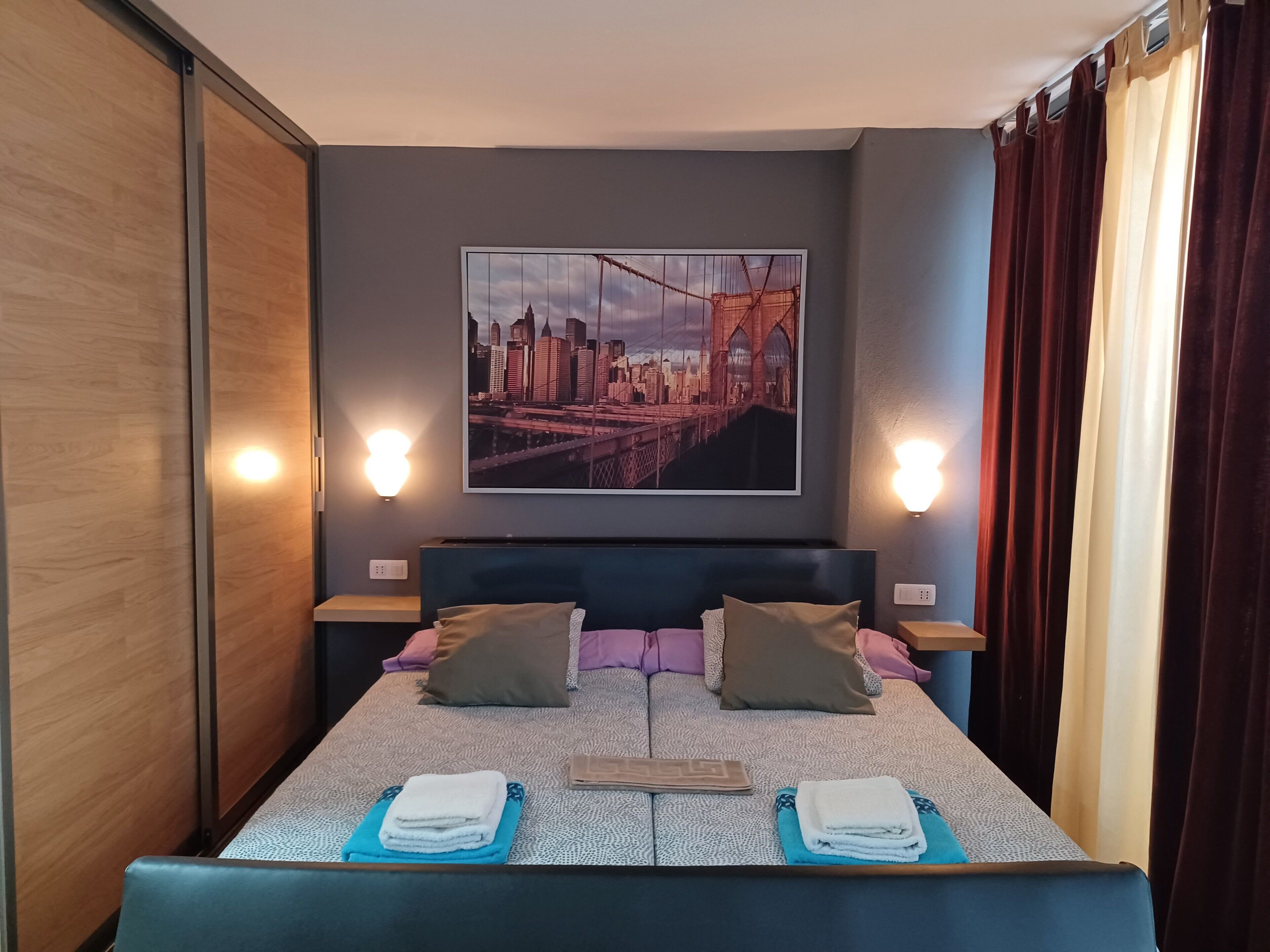 Een hotelkamer met een bed, kussens, een stadsgezicht schilderij, nachtlampjes en gordijnen.