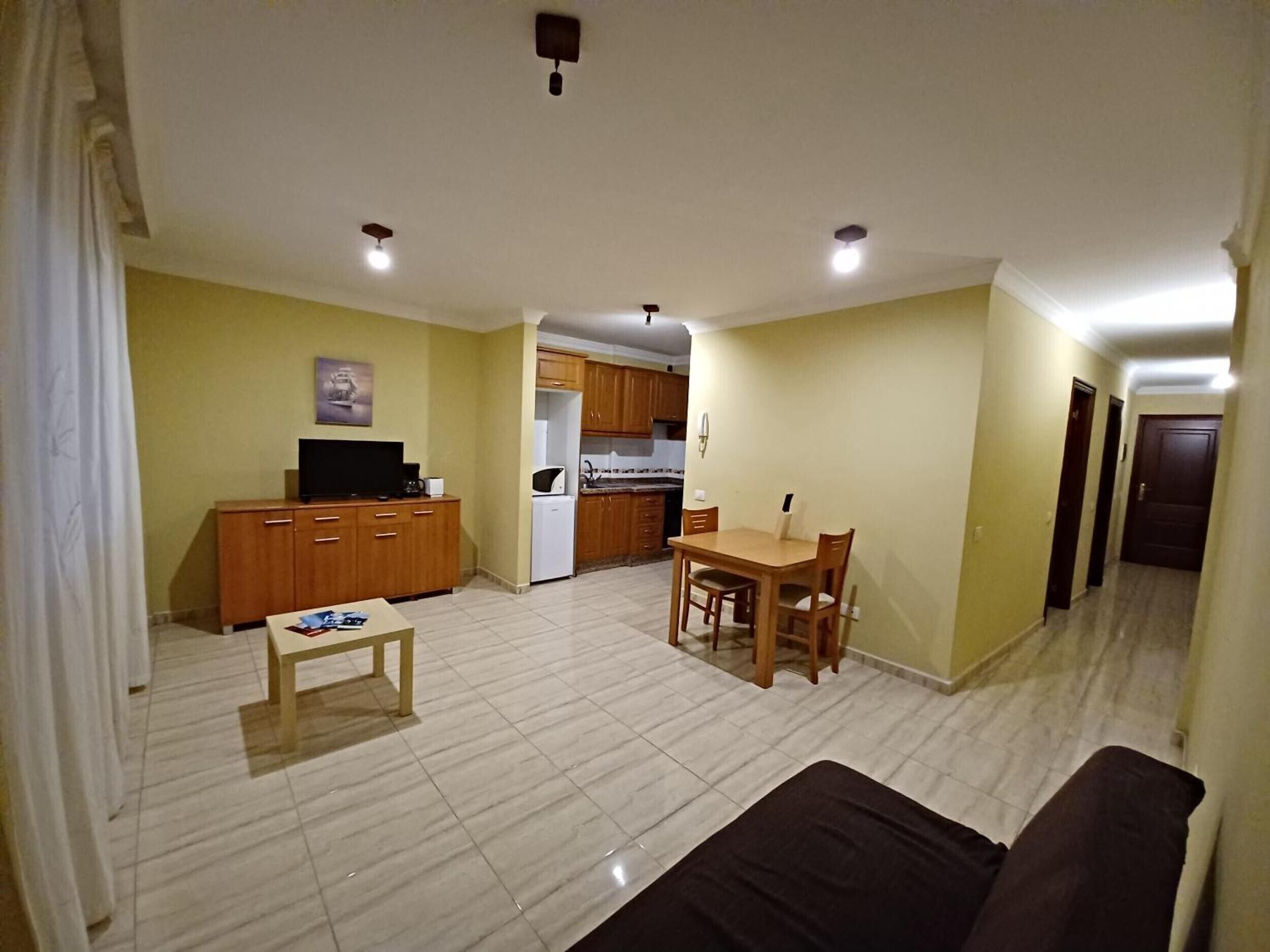 A living room with a sofa, a coffee table, and a TV. There is a kitchen area with cabinets and a refrigerator.