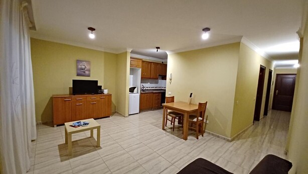 Sala de estar com sofá, mesa de centro e televisão. Há também uma área de cozinha com armários e geladeira.
