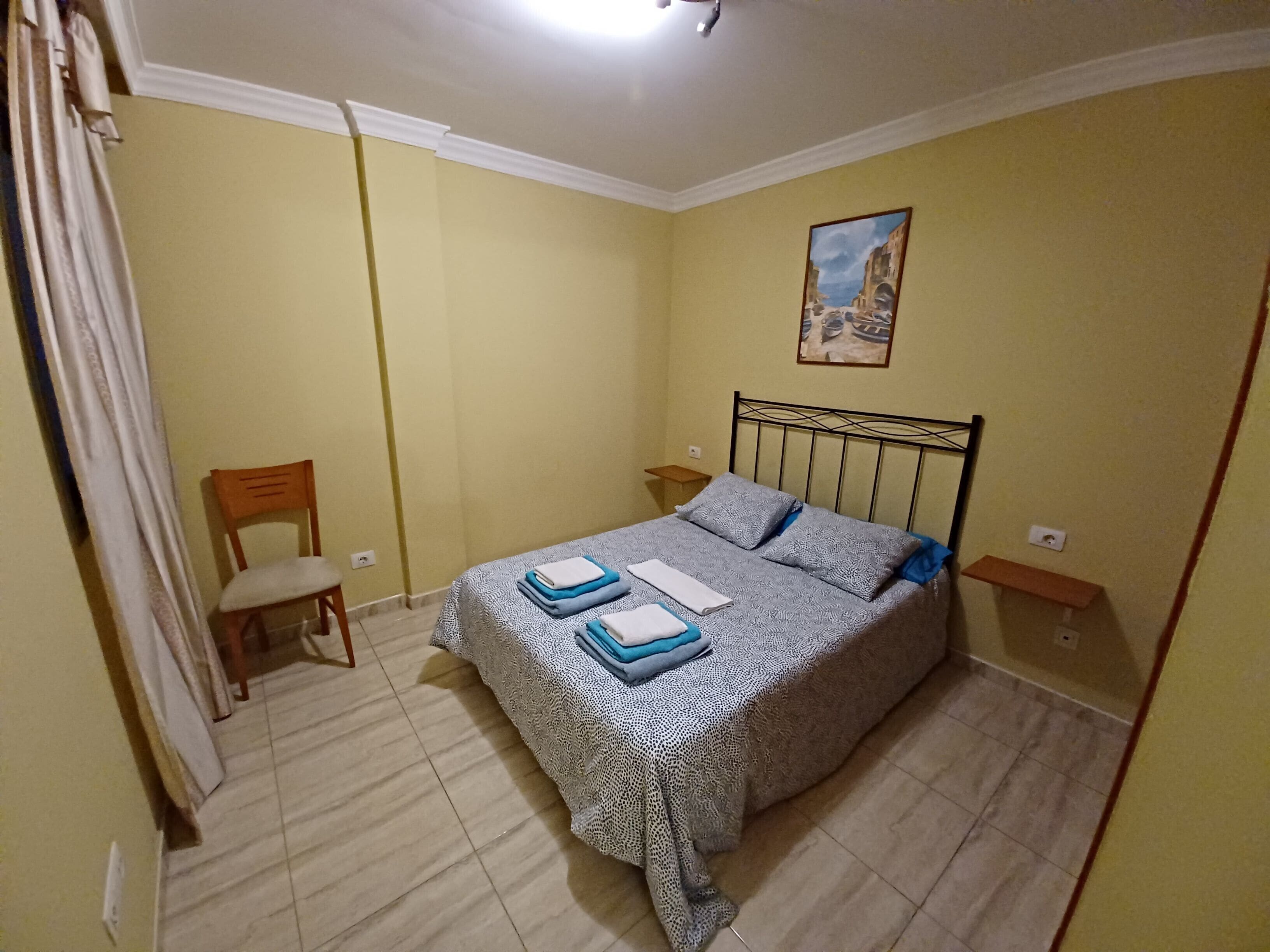 A bedroom with a bed, a chair, a painting on the wall, and a small table.