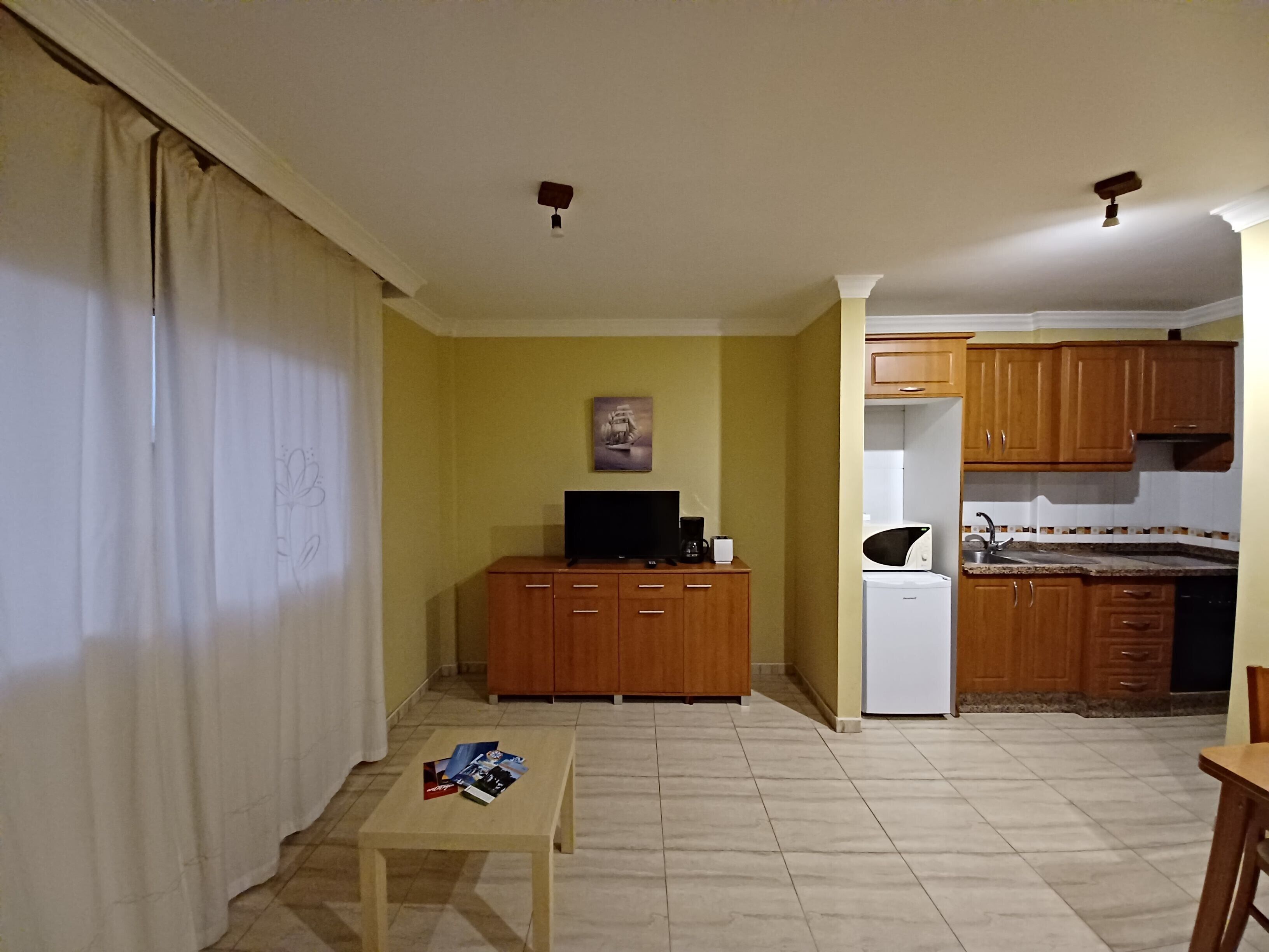 Een woonkamer met een televisie, een salontafel met tijdschriften, en een keukenhoek met houten kasten en een koelkast.