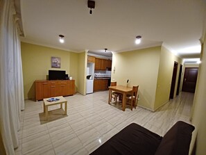 A living room with a sofa, a coffee table, and a TV. There is a kitchen area with cabinets and a refrigerator. The room has a tiled floor and a door leading to another room.