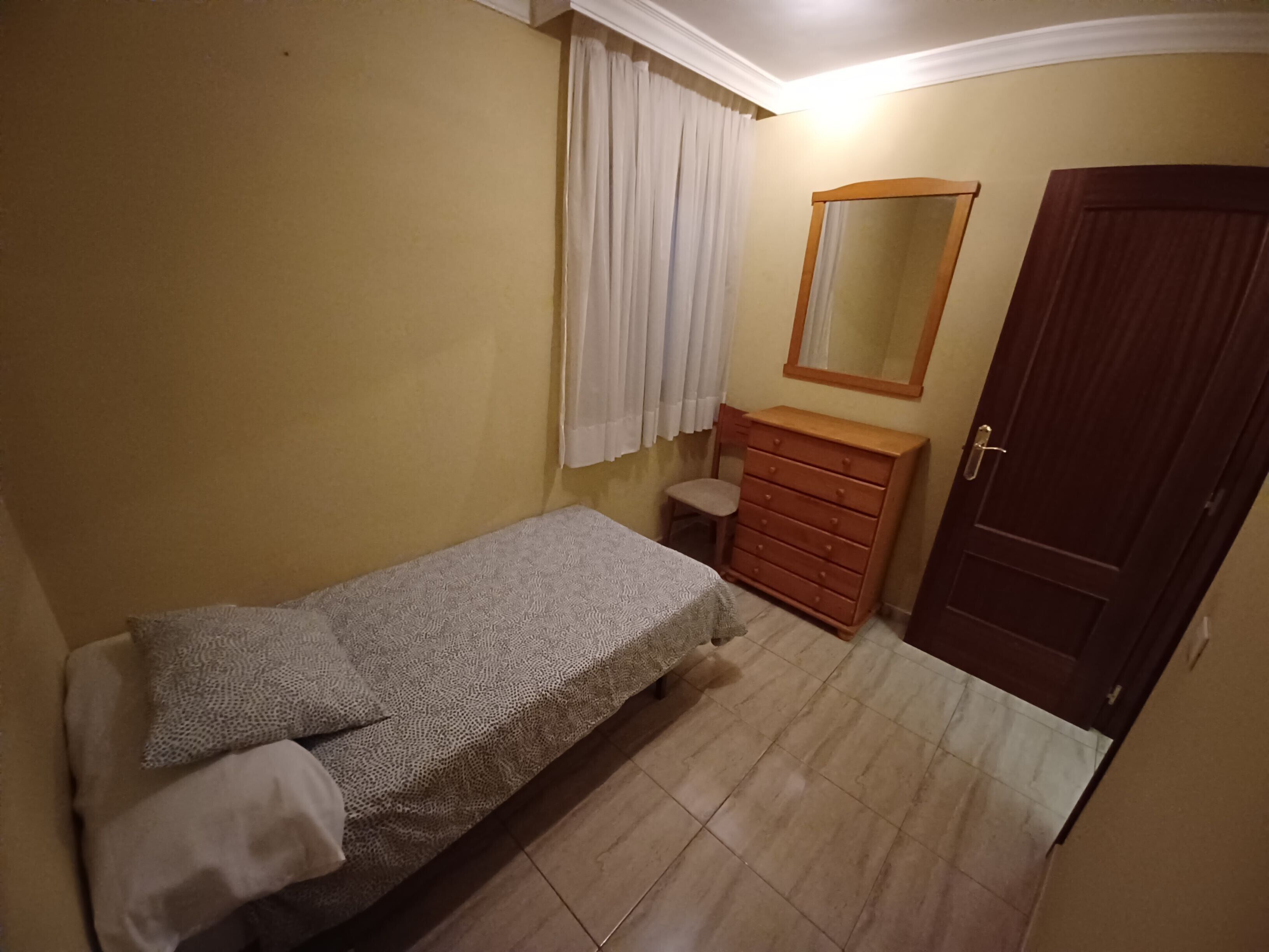 Een slaapkamer met een enkel bed, een houten dressoir, een spiegel en een deur.