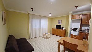 Um espaço de estar compacto com sofá, uma mesinha, televisão em um móvel e uma pequena cozinha.