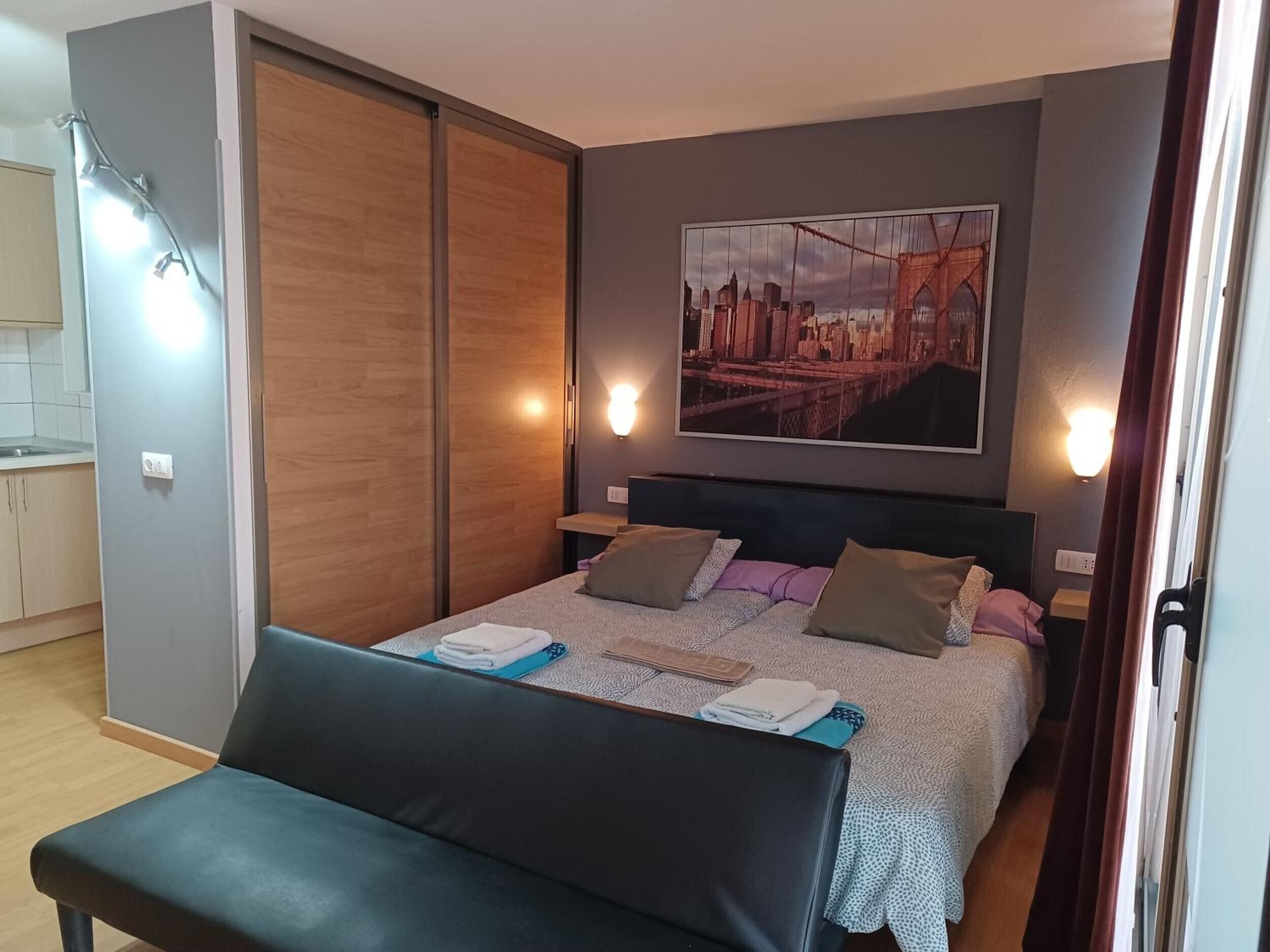 Een slaapkamer met een bed, een bankje, een stadsgezicht schilderij en een schuifdeur.