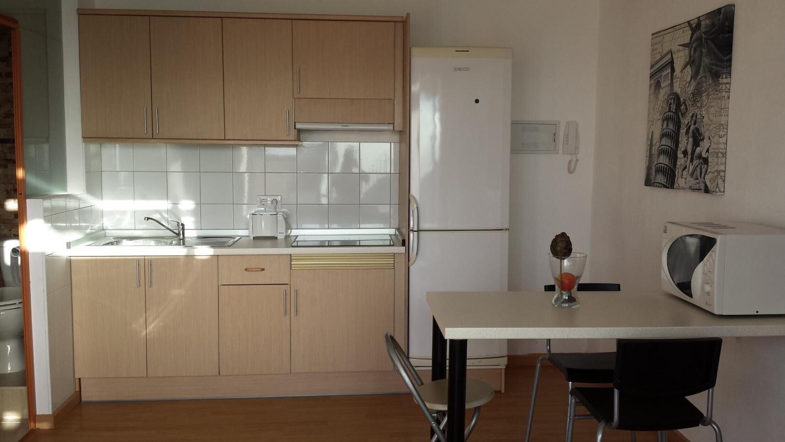 Una cuina compacta amb armaris de fusta, un refrigerador blanc, un microones i una taula de dinar petita amb una cadira.