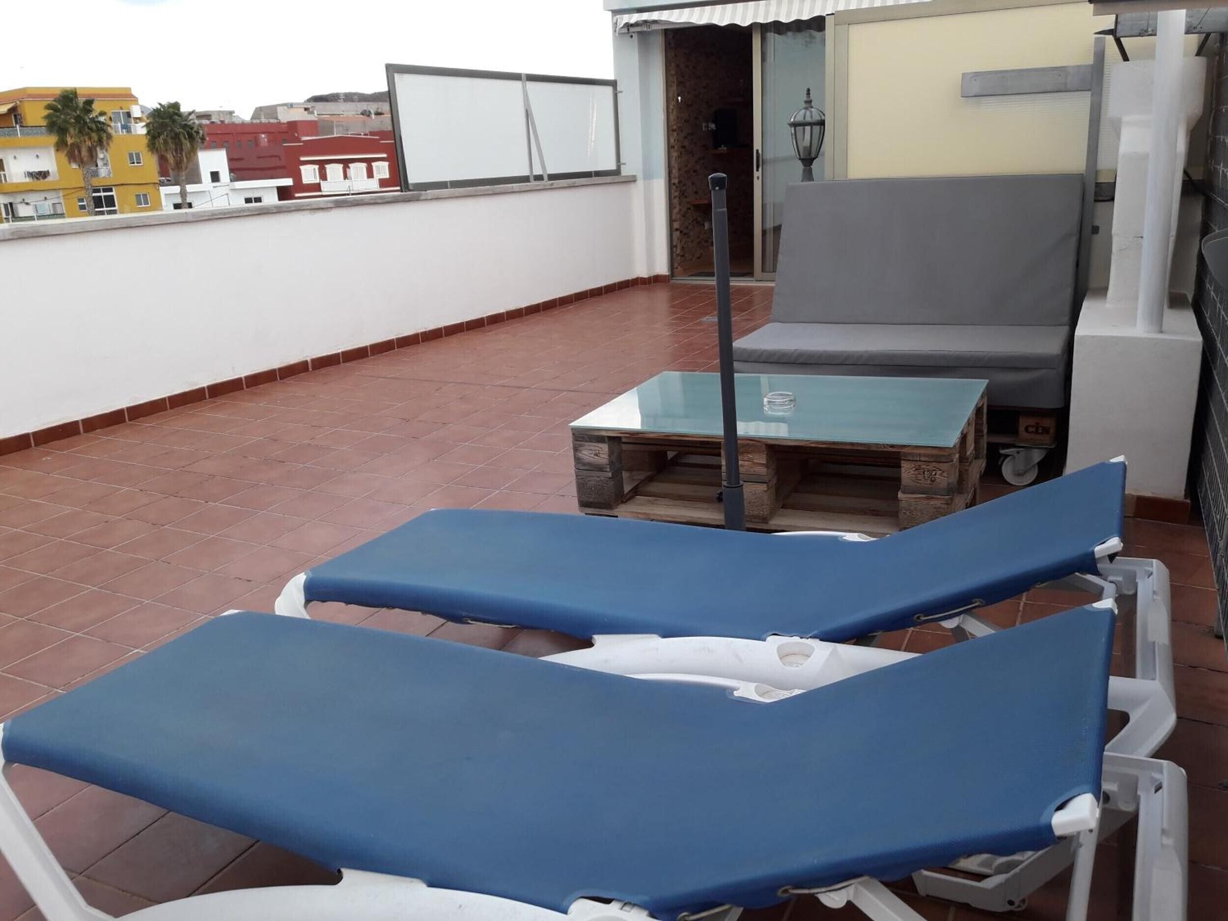 A rooftop terrace with two blue lounge chairs, a glass table, and a view of buildings in the background.