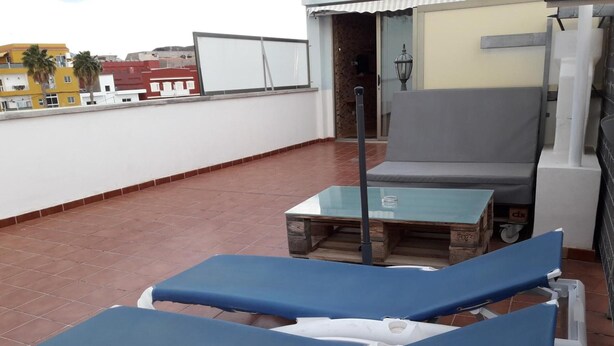 Um terraço no telhado com duas espreguiçadeiras azuis, uma mesa de vidro e vista para edifícios ao fundo.