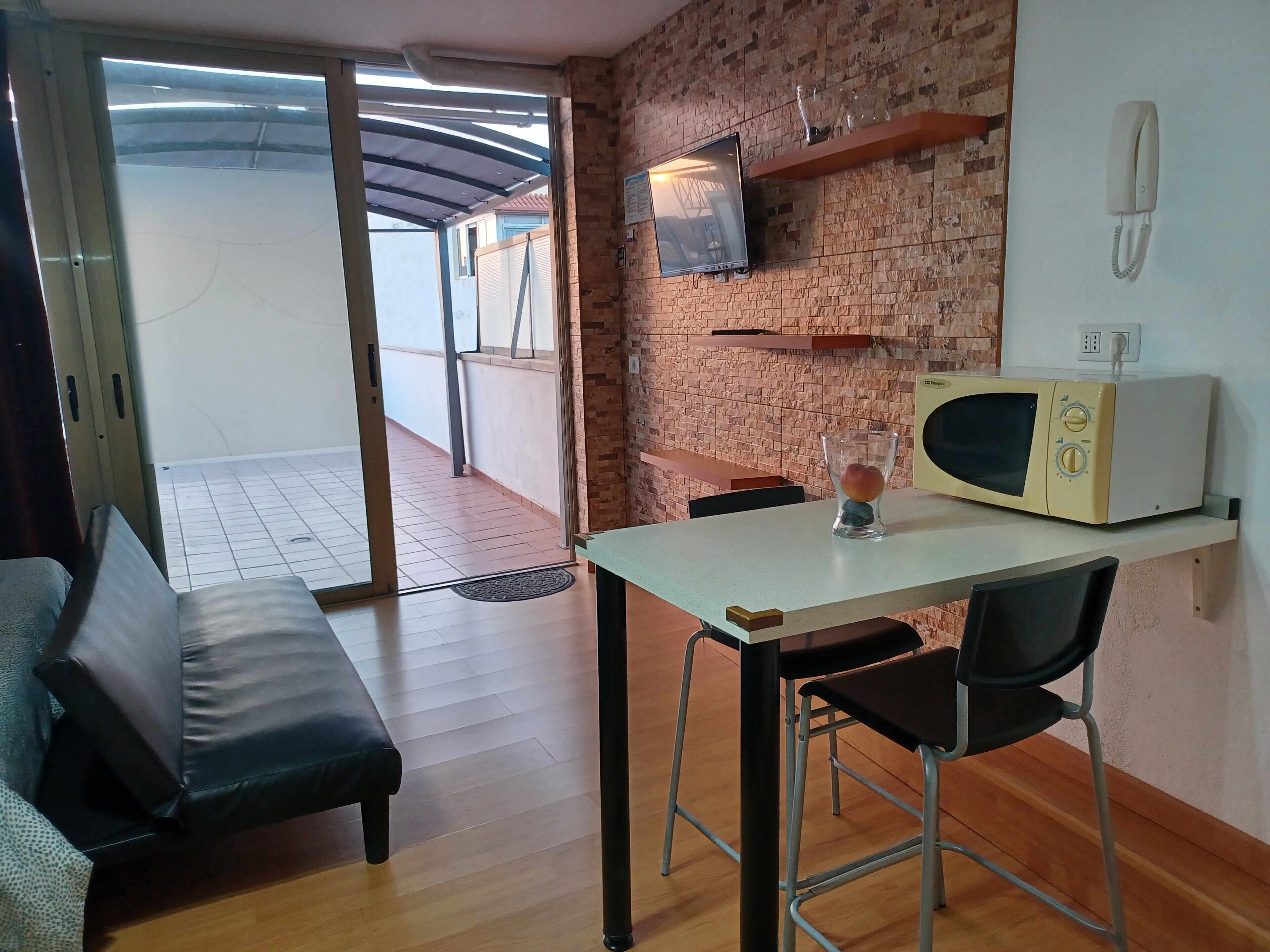 Een kamer met een bank, een tafel met een stoel, een magnetron, een televisie en een bakstenen muur.