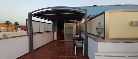 Un balcon extérieur couvert, équipé d’une table et de chaises, d’une lampe, et offrant une vue sur les bâtiments en arrière-plan.