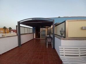 Een overdekt balkon in de open lucht, met een tafel, stoelen, een lamp en een uitzicht op gebouwen in de verte.