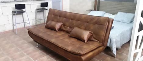 Un canapé en cuir marron avec quatre coussins, dans une pièce dont le sol est recouvert de carreaux, avec deux chaises en métal et un lit recouvert d’une couvre-lit à motifs.