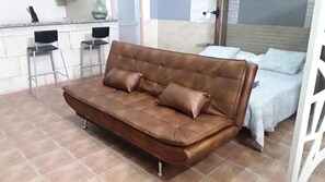 A brown leather sofa with four cushions in a room with tiled flooring, two metal chairs, and a bed with a patterned bedspread.