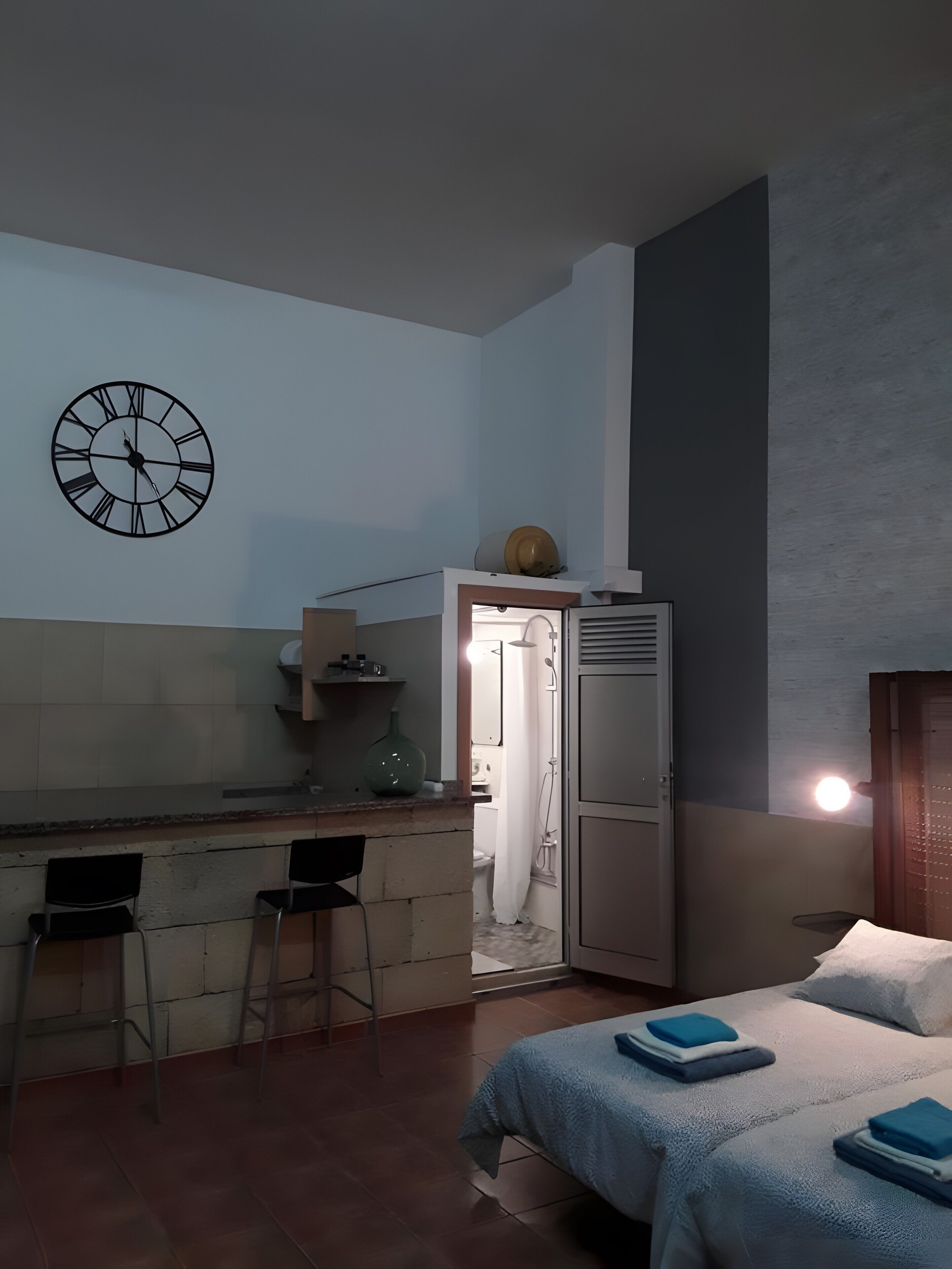 Une petite pièce comprenant un coin cuisine, un lit avec des serviettes soigneusement pliées, et une horloge murale.