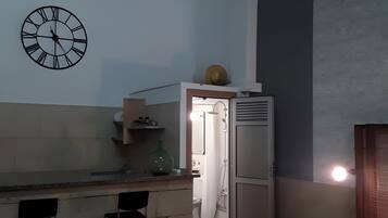 Une petite pièce comprenant un coin cuisine, un lit avec des serviettes soigneusement pliées, et une horloge murale.