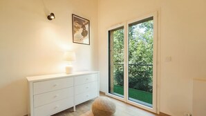 Une chambre moderne avec une commode blanche, une lampe, un pouf rond et une grande porte-fenêtre coulissante donnant sur un espace extérieur verdoyant.