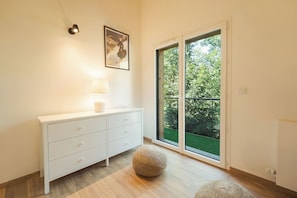 A modern room with a white dresser, a lamp, a round pouf, and a large sliding glass door leading to a green outdoor area. - Cimes - Modern, Sleeps 8 | New | Family-Friendly | Quiet Location (Chamonix)