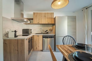 A compact kitchen with wooden cabinets, a stainless steel dishwasher, and a dining area with a wooden table and chairs. - Cimes - Modern, Sleeps 8 | New | Family-Friendly | Quiet Location (Chamonix)