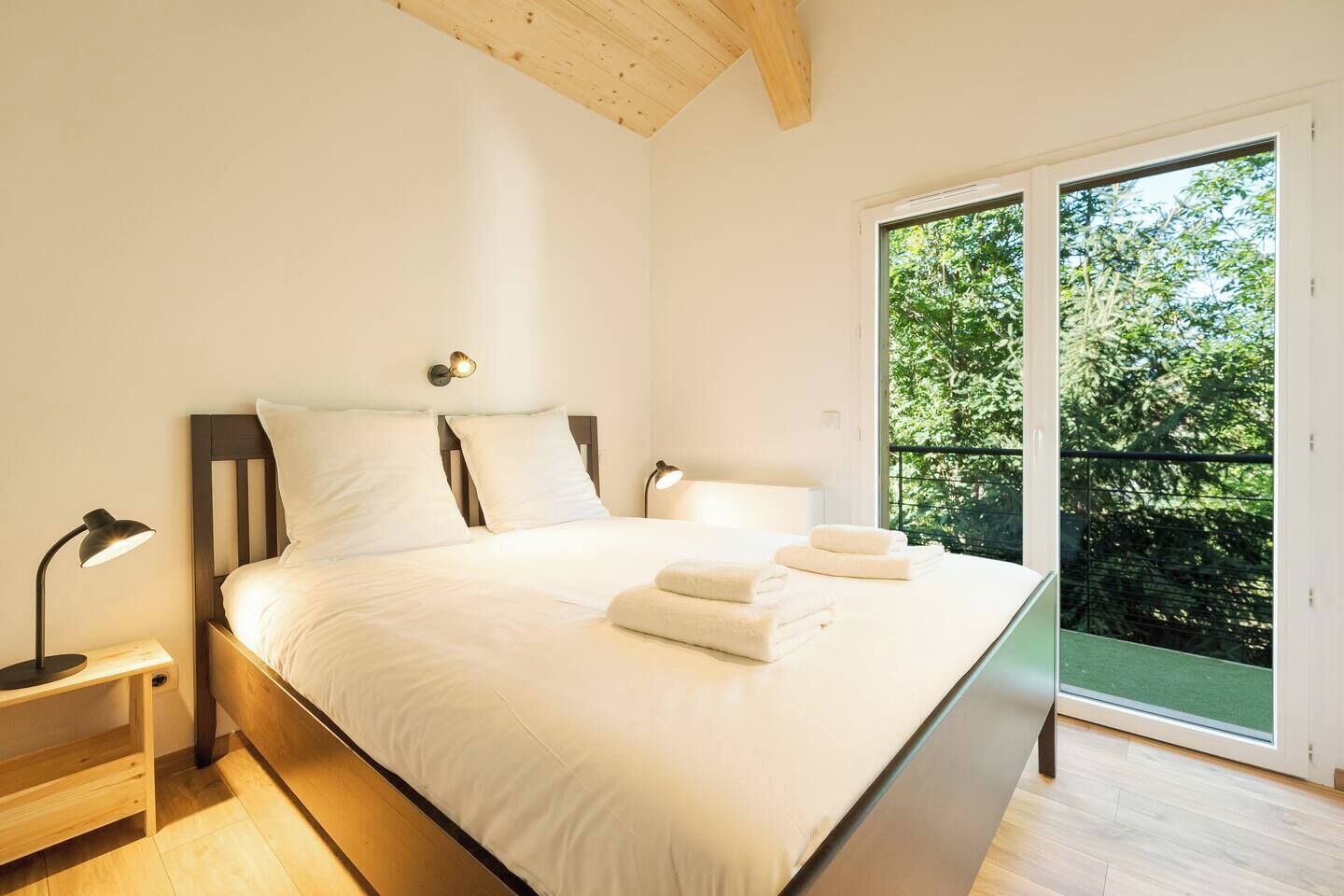 A bedroom with a large bed, bedside lamps, and a view of greenery through a sliding glass door.
